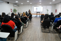 Várias pessoas em um auditório. Elas estão sentadas e assistem uma pessoas que está de pé que falando, trajada de roupa escura.