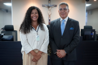 Mulher parda de cabelos longos cacheados ela veste roupa clara e está ao lado de um homem alto de meia idade que usa óculos e  está trajado de terno escuro.