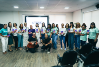 Diversas mulheres, todas de frente, usando camisas brancas, calças jeans e de tecido, à frente elas, uma mulher e um homem agachados usando calças e blusas pretas,. atrás de todos eles, um projeto branco com o nome CTESK escrito em letras azuis claras.