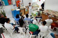 Várias pessoas sentadas em cadeiras brancas assistem uma mulher de avental branco e toca preta que está de pé falando.