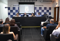 A imagem retrata uma reunião realizada em um auditório, onde quatro pessoas estão sentadas à mesa principal, conduzindo a discussão. Ao fundo, um telão exibe a imagem de um participante remoto. O auditório conta com diversos espectadores sentados em cadeiras pretas, atentos ao debate. A identidade visual no painel de fundo sugere um ambiente institucional ou corporativo.