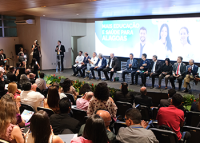 A imagem mostra um evento em um auditório, com um grande público acompanhando uma solenidade. No palco, diversas autoridades estão sentadas em uma mesa de honra, enquanto uma pessoa discursa em pé ao lado. Ao fundo, um telão exibe a mensagem "Mais Educação e Saúde para Alagoas", indicando o tema da cerimônia. O ambiente está decorado com arranjos florais na frente do palco, e a plateia acompanha atentamente.