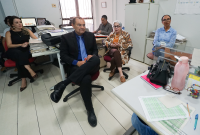 Há três mulheres e dois homens sentados, em uma sala de piso e paredes na cor branca. O mobiliário do ambiente é cinza claro.