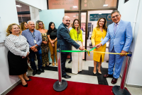 A imagem mostra um grupo de pessoas participando de uma cerimônia de inauguração. No centro da foto, três indivíduos seguram uma fita verde e amarela prestes a ser cortada. Um homem de terno escuro e gravata sorri enquanto segura a fita junto a uma mulher vestida de amarelo e um homem de terno azul claro. Atrás deles, outras pessoas observam o momento, algumas sorrindo. O ambiente parece ser o interior de um prédio institucional, com uma placa de vidro ao fundo indicando a "Central de Atendimento". 