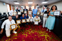 A imagem mostra um grupo de pessoas reunidas em um auditório ou plenário, posando para uma foto de premiação. Todos seguram placas com a inscrição "Mérito 2024" e demonstram expressões de felicidade e orgulho. O ambiente tem uma decoração formal, com um crucifixo ao fundo e cadeiras organizadas no espaço. O chão está coberto por confetes dourados, sugerindo uma celebração recente. Os premiados estão organizados em duas fileiras, alguns em pé e outros agachados ou ajoelhados na frente, exibindo suas placas 