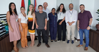 A foto mostra um grupo de nove pessoas reunidas em um ambiente institucional. Todos estão sorrindo e posando para a foto, vestindo trajes formais e casuais. Ao fundo, há bandeiras com laços decorativos, sugerindo um ambiente oficial. O grupo é diverso, composto por homens e mulheres, e aparenta estar em um momento de confraternização ou registro de um evento especial.