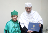 A imagem mostra duas pessoas vestindo trajes acadêmicos formais em uma cerimônia da Universidade Federal de Alagoas (UFAL). A mulher à esquerda veste uma beca verde com detalhes ornamentados e um chapéu correspondente, enquanto o homem  à direita usa uma beca branca e segura um diploma com o brasão da UFAL. O ambiente sugere um evento oficial de outorga de título ou formatura.