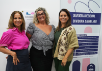 Três mulheres brancas sendo duas loiras e uma de cabelo castanho em frente a uma placa com o nome ouvidoria regional e ouvidoria da mulher 
