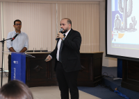 Homem branco, calvo, de paletó preto e camisa branca em pé apresentnado o projeto; ao lado direito dele, o mestre de cerimônia, também em pé, de camisa social branca 