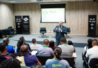 Há um homem de terno cinza falando de pé para uma plateia senda em um auditório