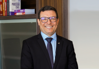 Homem de pele clara, cabelos curtos e escuros, usando óculos e sorrindo. Ele veste terno preto, camisa azul clara e gravata azul escura com pequenos pontos. Ao fundo, há uma estante com livros jurídicos.
