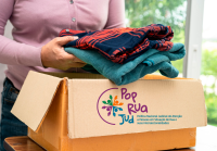 Pessoa coloca roupas dobradas em uma caixa de papelão com a logomarca do Pop Rua Jud. A caixa está sobre uma mesa e, ao fundo, há uma janela com vista desfocada.