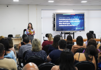 Auditório com várias pessoas sentadas, assistindo a uma palestra. Uma mulher está em pé, falando ao microfone, ao lado de uma tela grande que exibe o tema: “Fortalecendo a Justiça, com saúde e prevenção das doenças osteomusculares”.