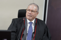 Homem branco, de cabelo grisalho, de paletó preto, blusa branca e gravata azul sentado.