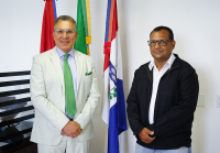 Há duas pessoas em pé, lado a lado, em um ambiente interno. Ao fundo, há três bandeiras. À esquerda, está um homem de cabelos curtos grisalhos e óculos. Ele veste um terno verde-claro com gravata e lenço de bolso em um tom de verde mais vibrante, além de camisa clara. Está sorrindo e com as mãos cruzadas à frente do corpo. À direita, está um homem, também usando óculos. Ele veste camisa branca, jaqueta preta e calça jeans clara. Está com expressão neutra e também com as mãos cruzadas à frente do corpo.