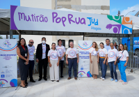 Há 11 pessoas em pé, sorrindo, em frente a uma tenda com uma grande faixa escrita "Mutirão Pop Rua Jud Alagoas". A maioria das pessoas veste camisetas brancas do evento, com o logotipo colorido do “Pop Rua Jud” no centro. Duas mulheres estão com roupas diferentes.