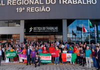 A imagem mostra um grande grupo de pessoas reunido em frente ao prédio do Tribunal Regional do Trabalho da 19ª Região (TRT-AL), em Maceió. Participantes seguram bandeiras de sindicatos e movimentos sociais, como CUT, FETAG-AL, SINTEAL e outros, em um ato de mobilização coletiva. Ao fundo, vê-se a fachada do Tribunal com bandeiras hasteadas.