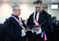 Dois magistrados estão em destaque, trajando togas pretas com detalhes em renda branca nos punhos e no colarinho, além de faixas nas cores azul, branca e vermelha, atravessadas no peito, e medalhas penduradas. Eles estão em pé dentro de uma sala de sessões, em um ambiente institucional, olhando atentamente para um documento azul que seguram juntos