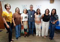 A imagem mostra sete pessoas, sendo seis mulheres e um homem, todas em pé e sorrindo para a câmera. Elas estão lado a lado, em ambiente interno, na sala institucional, com piso de madeira e paredes brancas. No fundo, há duas placas fixadas na parede e cadeiras azuis encostadas. As pessoas vestem roupas casuais, com cores variadas. O homem, que está ao centro, usa camisa azul escura e calça jeans, com crachá no pescoço. 
