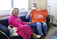 Mulher branca de cabelos loiros usando vestido rosa sentada em um sofá à esquerda; à direita, homem de paletó azul claro sentado segurando uma camisa de cor laranja