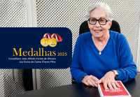 À direita da imagem, uma mulher, de cabelos curtos e brancos, veste uma blusa azul e usa óculos de armação escura. Ela está sorrindo e sentada à mesa, com as mãos apoiadas sobre um livro de capa vermelha. Usa um colar dourado e um relógio no pulso esquerdo. À esquerda da imagem, há um cartaz com fundo azul escuro.