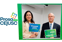 A imagem mostra duas pessoas posando sorridentes em uma moldura verde com cartazes nas mãos, durante o evento “Prosa com CEJUSC”. À esquerda, uma mulher segura o cartaz com a frase “Prosear para conciliar”, e à direita, um homem com traje formal segura outro com a frase “Com prosa a gente se entende”. Ao fundo, o ambiente interno reforça o clima institucional do evento promovido pelo CEJUSC do TRT da 19ª Região.
