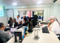 Em uma sala de audiências, cerca de dez pessoas estão reunidas em torno de uma mesa com computadores. À esquerda, um homem apresenta algo em um notebook enquanto os demais assistem com atenção. À direita, uma mulher de cabelos brancos e roupa clara observa atentamente a apresentação. Livros jurídicos.