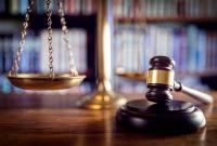 A imagem mostra uma composição simbólica do sistema judiciário: em primeiro plano, há um martelo de juiz (malhete) sobre uma superfície de madeira, indicando a autoridade das decisões judiciais. Ao fundo, levemente desfocada, aparece uma balança dourada da justiça, representando o equilíbrio e a imparcialidade do Direito. Estantes com livros jurídicos completam o cenário, reforçando o contexto de um ambiente jurídico ou tribunal.