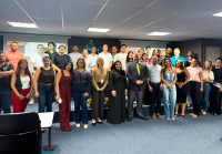 Fotografia mostra um grupo de pessoas reunidas em um auditório para uma foto coletiva. Elas estão dispostas em duas fileiras: a primeira em pé no chão do auditório e a segunda em pé sobre um pequeno palco. Ao fundo, há dois telões de projeção exibindo slides com rostos de outras pessoas, sugerindo uma apresentação.