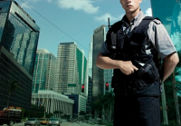 A imagem mostra um agente de segurança patrimonial de uniforme e colete tático em pé, com as mãos próximas à cintura, diante de um cenário urbano composto por prédios comerciais espelhados e trânsito ao fundo. A composição transmite uma sensação de vigilância e proteção em área corporativa.