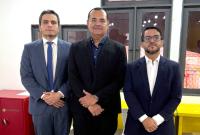 Três magistrados posam juntos para foto em ambiente interno, vestindo ternos formais. Ao fundo, janelas com cortinas translúcidas e mobiliário escolar.