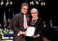 Foto de homem homem alto à direita usando toga preta e mulher idosa , à esquerda, de cabelo grisalho  usando vestido preto; ambos estão segurando um certificado branco com bordas azuis.