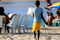 Menino negro de costas na areia de uma praia voltado para o mar, usando bermuda amarela e uma blusa azul de listras azuis escuros e segurando um depósito de comida na mão esquerda e, na direita, uma churrasqueira