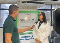Ao lado direito, mulher morena de cabelo preto e sobretudo cor de creme em pé concedendo entrevista; ao lado esquerdo, o repórter usando blusa verde e segurando o microfone na mão esquerda