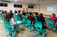 À esquerda, à frente do público sentado, uma mulher usando calça preta e blusa cinza ministra palestra em pé.