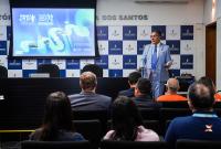 Homem alto de terno azul claro à frente do público do lado direito; à direita dele, um telão com o fundo azul claro