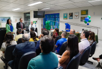 Mulher de blusa verde clara à esquerda  e dois homens em pé à direita; à frente dele, várias pessoas sentadas