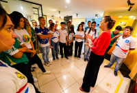 Há várias jovens de uniformes escolares em pé em um cículo. No centro,  há uma mulher de blusa vermelha e calça preta falando para esses jovens.