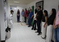 Um homem magro, moreno, de roupa branca e chapéu preto ao lado esquerdo fazendo uma apresentação para vários servidores em pé ao seu lado direito