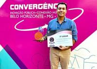 Fundo rosa com letra branca escrito convergência e um homem branco com camisa jeans azul clara com um certificado na mão