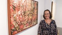 foto de mulher branca de olhos verdes e blusa preta ao lado de um quadro de manguezal