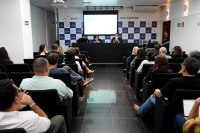 Dois homens sentados ao centro do birô, atrás deles, um telão; à frente deles, várias pessoas sentadas