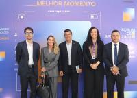 Cinco pessoas, sendo duas mulheres e três homens, em pé em pose para a foto. Os homens estão de ternos escuros e as mulheres estão vestindo roupas sociais; atrás deles, um banner grande de fundo azul