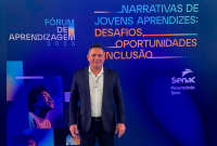 Homem moreno de blaser escuro e camisa branca em pé; atrás dele, um gande banner azul