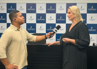 Entrevista em ambiente interno: repórter da TV Gazeta, homem pardo de cabelo curto e escuro, entrevista a juíza Estefânia Fernandes, mulher branca, loira, de cabelos lisos e médios, diante de painel do TRT-AL. Ambos estão de frente um para o outro em posição formal.