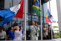 A imagem mostra um grupo de pessoas participando de um ato simbólico de hasteamento de bandeiras em frente a um prédio com fachada de vidro. Três pessoas estão erguendo bandeiras em mastros: uma mulher loira com blusa branca de bolinhas pretas levanta a bandeira do estado de Alagoas (nas cores azul, branca e vermelha); ao lado, um homem de terno claro e gravata azul levanta a bandeira do Brasil; e outro homem com paletó bege ergue a bandeira do orgulho LGBTQIAPN+.