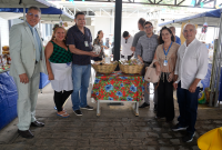 A imagem mostra um grupo de seis pessoas posando sorridentes em frente a uma mesa com toalha colorida e cestos contendo produtos variados. Ao fundo, há barracas de feira e outras pessoas circulando pelo local, que está decorado com bandeirolas típicas de festas juninas. O ambiente é coberto e tem clima festivo.