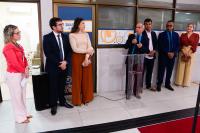 Há sete pessoas em pé, alinhadas contra uma parede interna, participando de um evento. Uma delas — o quarto da direita para a esquerda — segura um microfone e está falando ao público. O ambiente é institucional, com piso claro, um tapete vermelho à frente e painéis decorativos ao fundo. As pessoas estão vestidas de forma formal, sugerindo uma cerimônia ou abertura de exposição
