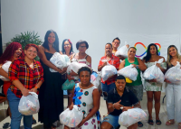 Grupo de pessoas posa sorridente para foto segurando sacolas de doações. Ao fundo, há uma parede branca com um cartaz contendo um símbolo colorido.