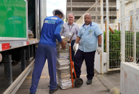 Três homens altos ao lado de um caminhão com um baú verde; no meio deles, um carrinho com duas rodas cheio de processos